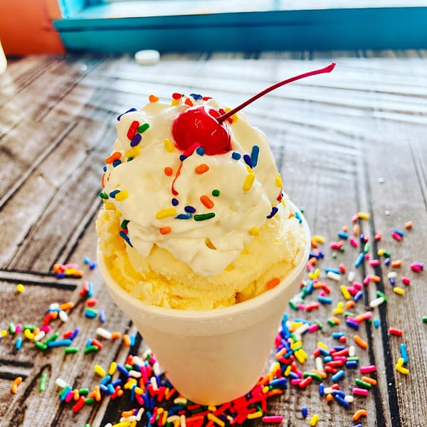 Vanilla ice cream in a white cup, topped with whipped cream, sprinkles, and a cherry.