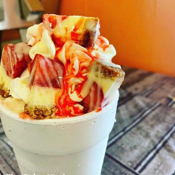 A white cup filled with cheesecake chunks, strawberries, whipped cream, and red and white drizzles.