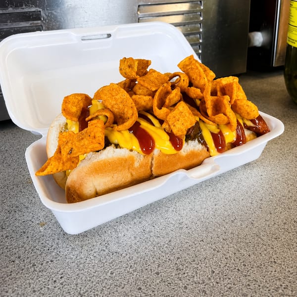 Two hot dogs topped with chili, cheese sauce, ketchup, and crunchy chips in a white takeout container.
