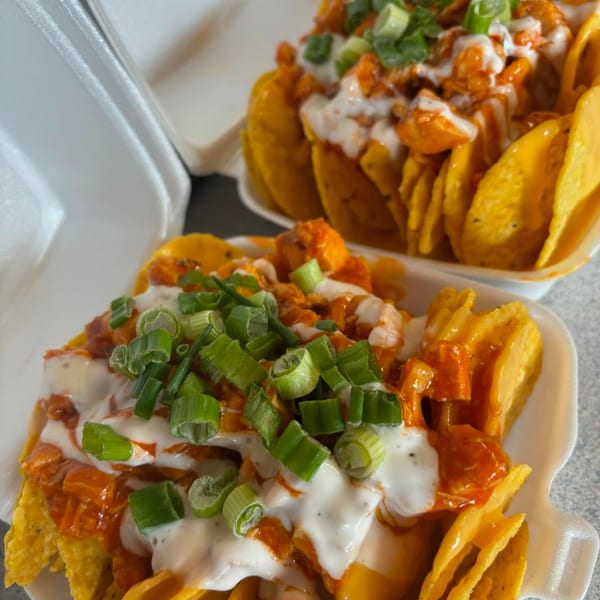 Two takeout containers filled with nachos topped with buffalo chicken, ranch, and green onions.